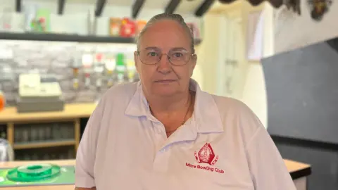 Jack Maclean/BBC A woman standing inside a building in front of a bar. She is looking directly at the camera with a serious look on her face. She is wearing a white short-sleeved polo shirt which says "Mitre Bowling Club" on it, with the club's emblem in red stitching. She is wearing glasses. Her grey hair has been pulled back.