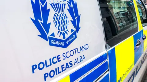 A close up of the side of a police van parked in a street. The van has Police Scotland in English and Gaelic. The vehicle is white with blue and yellow markings.
