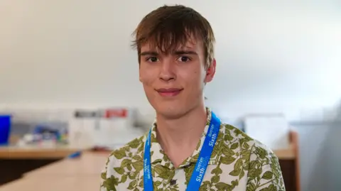 Martin Giles/BBC A 17-year-old wearing a cream and green patterned shirt. He has a blue lanyard round his neck. He has brown hair and is smiling at the camera.