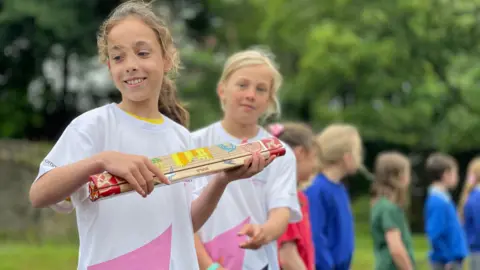 A girl wearing a white t-shirt, with wavy dark blonde hair tied back, is holding the wooden baton, which is colourfully painted. There are other students in a line to the right, blurred, stood outside with trees behind them.
