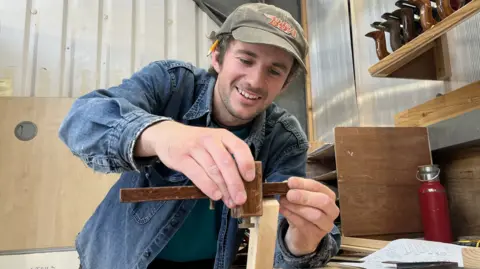 A man in a blue denim shirt marks a piece of wood using a tool. He is smiling. He is also wearing a green baseball cap with the BSA logo on it and has a yellow pencil tucked behind his right ear.