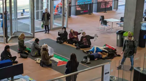 Protesters with signs and flags sit and lie on the floor of a modern building with a stone floor. 