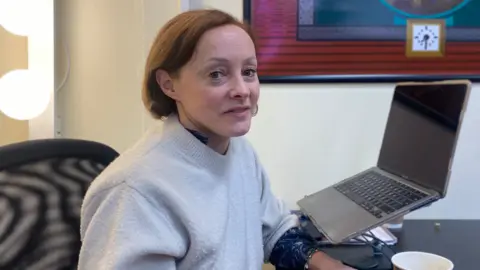 Mimi Turtle sits on an office chair at a desk with a laptop in front of her. She has a cream jumper on and brown hair. There are lights and artwork behind her.