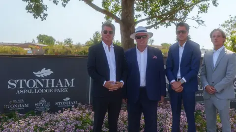 Indivisuals/Matt Filby Four men wearing three blue suits and a pale grey suit and white open neck shirts are standing next to a sign that reads Stonham Aspal Lodge Park. They are in front of a bed of pink Sweet William flowers. The three in blue suits are also wearing sunglasses, while one of them is wearing a white Panama hat.