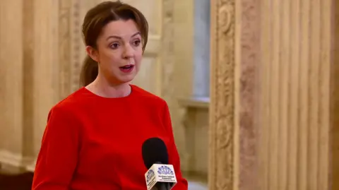 Liz Kimmins, wearing a red outfit and standing in front of microphone. She has brown hair pulled back into a ponytail and is standing in the Great Hall at Stormont.