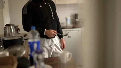 Asylum seeker wearing sportswear - a black top and white jogging bottoms - holds a spoon as he cooks in the kitchen.