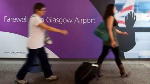 Two people walking through an airport, carrying suitcases. A sign behind them reads Farewell from Glasgow Airport.