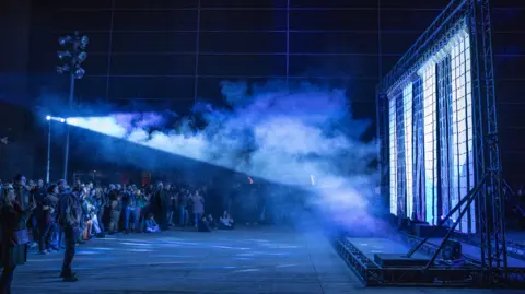 Parker Heyl A large curtain of tiles in a dark cavernous space underground. There is a crowd of people watching the installation move, as a projector shines blue and purple light across the room. The space is filled with smoke and scattered light.