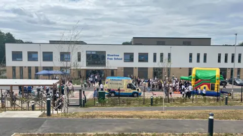 Nicola Haseler/BBC A new two-storey school called Rivertree Free School. There is a bouncy castle and an ice cream van outside and several pupils and adults. There is a path in the foreground.