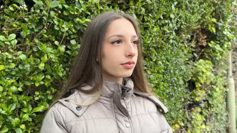 Image shows a student called Megan. She has long brown hair. The ends are blond. She is wearing a beige puffer coat which is zipped up. She has a neutral expression and is looking off to the right of the image towards the reporter, who is asking her questions. Behind her, the whole background is filled with green from a large green bush. 