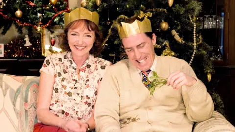 Melanie Walters and her co-star Rob Brydon sitting on a sofa during the filming of a Christmas episode of Gavin and Stacey. She is wearing a brown and pink flowery blouse, red skirt and a gold paper crown. Rob is sitting beside her wearing a beige cardigan, gold paper crown, and holding a sprig of mistletoe. Behind them is a decorated Christmas tree with red and gold baubles and tinsel. They are both laughing.