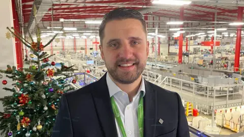 BBC/Phil Harrison Jay Brooks pictured overlooking the Medway Mail Centre. He is stood in front of a decorated Christmas tree, and is wearing a light blue shirt and a navy suit jacket. Jay is also wearing a green lanyard.