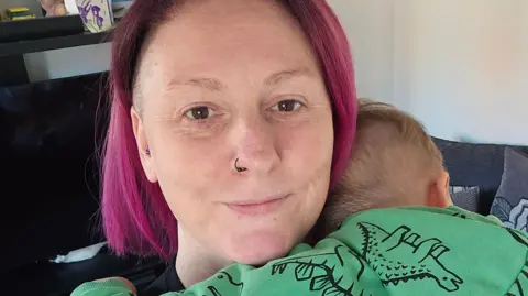 Kinship A woman with straight pink hair cuddles a toddler in a green hoodie with dinosaurs on.