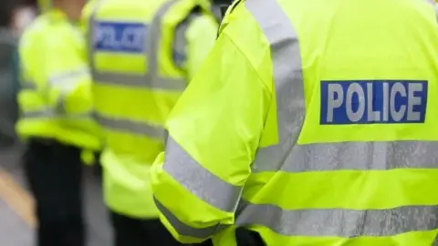Getty Images A police officer wears a luminous, high-visiblity jacket with POLICE written on the back in silver letters. Two other officers are visible in similar jackets in the background. 