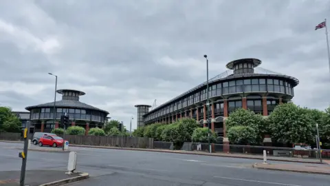 An image of an office block with a mainly glass upper floor in Nottingham. There is a similar building to the left. In the foreground is a wide road with several cars pulled up at traffic lights.