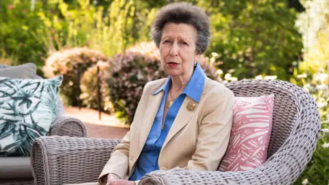 Her Royal Highness is sitting outside in a rattan garden seat. She is wearing a cream blazer with a blue shirt. She is also wearing a gold brooch. It is a sunny day with lots of greenery in the garden behind.