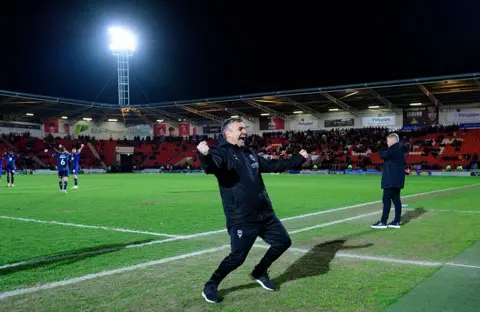 Chris Vaughan/Getty Images Michael Skubala, the head coach of Lincoln City, celebrates at the final whistle. He is wearing a black coat and tracksuit bottoms. Several players are in the background with their arms raised in celebration. They are standing on a green football pitch surrounded by football stadium seating. An illuminated floodlight is set against a dark sky.