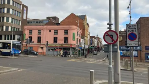 A road junction with a "no left turn" sign on the left. On the right a bus is about to turn into the road.