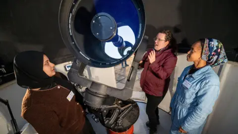 University of Leeds/Mark Bickerdike Three women are stood around a telescope, which is pointing towards the left of the image.