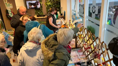 John Devine/BBC A small reception area inside a vets is packed with people looking at various goose merchandise.