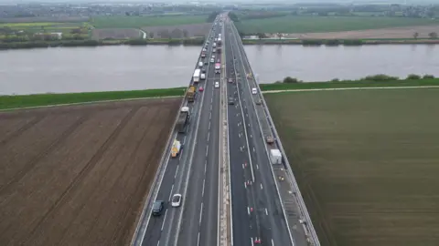 Aerial view of traffic making its way over the M62 Ouse Bridge. There are arable fields on either side and a stretch of water beyond.
