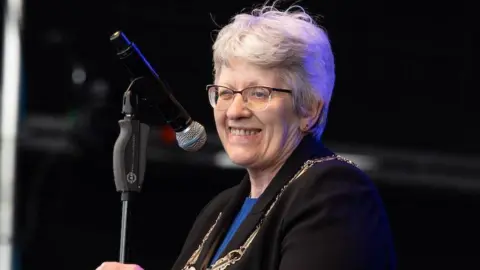 A woman with glasses and grey hair smiles in front of a microphone. She is wearing a black jacket with a blue T-shirt underneath and has a gold chain of office around her neck