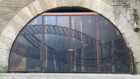 A railway archway with a brown metal frame inside it and drilled metal panels with some lettering and designs on it. 