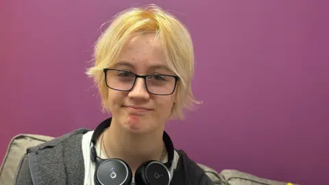 Robbie, a young boy with blonde hair, wearing black glasses. He is wearing a white t-shirt, grey zip up jacket alongside wearing black headphones which are below his neck.