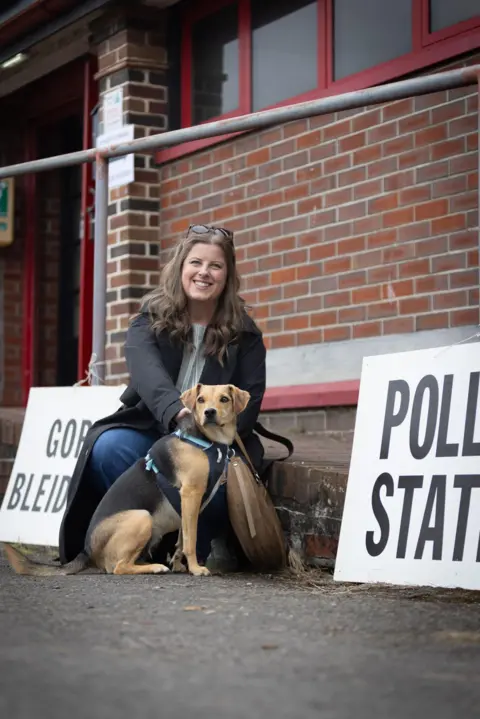 Ellen Cooper Brooke and her rescue dog Pheobe outside a polling station in Porthcawl. Phoebe is a black and tan dog with a chest harness on. Brooke is crouched down behind her and is wearing blue jeans with a black jacket. She has long wavy light brown hair and glasses resting on her head.
