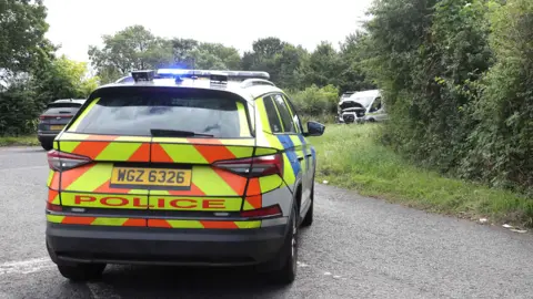 A police car with a car in the distance and a white van also in the distance. There is grass and trees along the side of the road.