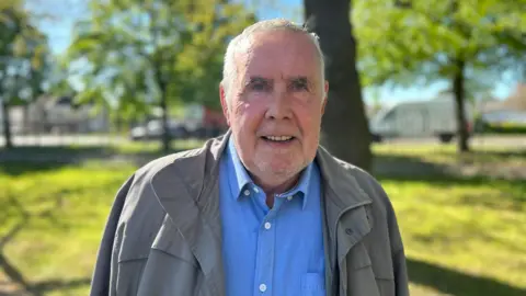 Head and shoulders image of John McKay. He has short white hair, stubble and is wearing a grey coat and blue shirt. He is standing in front of some trees and a patch of grass. 