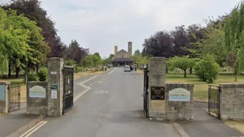 The entrance gates to Coney Hill Cemetery and Crematorium in Gloucester. Big stone posts with metal gates lead to a long driveway between green spaces with trees to the crematorium building.