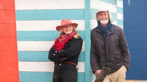 BBC/Tern Television/Kirsty Anderson Two people lean against a blue and white painted beach hut. The woman has blonde hair and wears a pink fedora hat, a red scarf and a dark coloured long coat. The man has a grey beard, a light coloured cap on, and a dark waterproof coat with beige trousers.