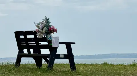 AndrewP An empty bench sits on grass with a view over water towards land in the far distance. A small yacht can be seen in full sail. A bunch of flowers with a note is attached to the back of the bench.