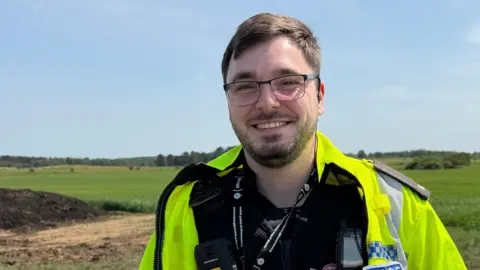 A man wearing a black police uniform and yellow hi-vis jacket smiles as he stands in a green field under a blue sky. He has short brown hair and a matching beard and wears glasses.