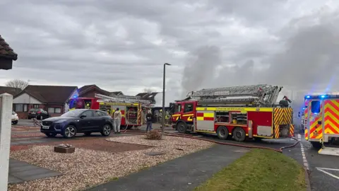 Fire engines and an ambulance gathered around a house with smoke billowing into the air