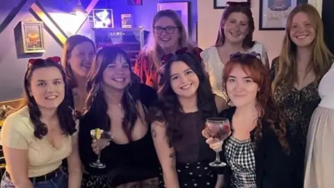 A group of young women pose together with their drinks in a pub and all smile.