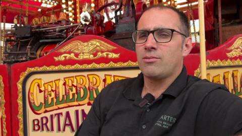 Joby Carter wearing a black top and glasses, standing in front of a colourful painted fairground ride