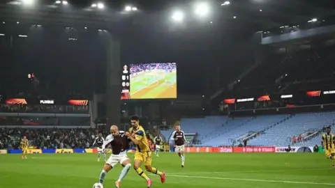 Getty Images An Aston Villa player is tackled by his Maccabi-Tel Aviv opponent. In the background there are other players running towards them, along with Villa Park stands where the crowd can be seen. 