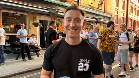 Danny Speck stood outside a pub, smiling at the camera. He is wearing a black T-shirt with white branding on. Behind him are groups of Hull City fans.