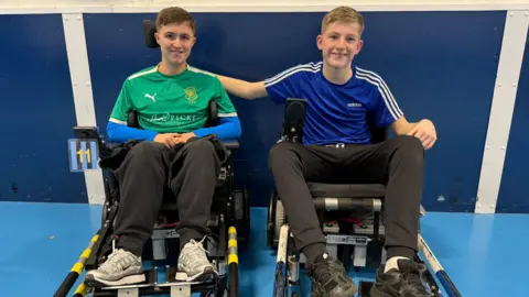 Two brothers sat in mechanical chairs inside. They're both smiling and the boy in the blue top has his arm round the other's shoulder. George, on the left, is in a green football shirt.