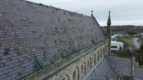 Aerial image of the slanted main roof of a church. We can see grey roof slates, some of which have clearly been displaced. The shot shows a full side profile of the church roof, with the front of the church towards the back of the imae. We an also see a landscape beyond the church - some building, a road and a hill.