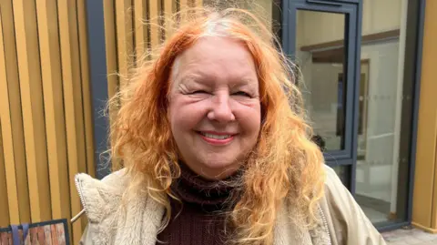 Catriona Goldhammer is standing in front of a wooden panelled building smiling at the camera and wearing a fur cream coat and maroon turtle neck jumper. She has orange wavy hair and red lipstick on. 