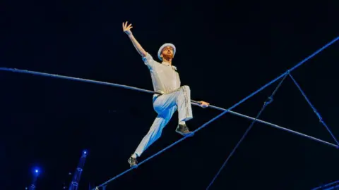 Chris Dempsey Ellis dressed in all white trousers and shirt, with a white hat, walks along a wire above a crowd. He wears glasses and has a ginger beard. He is holding a long balance pole above the crowd, with one hand outstretched. 