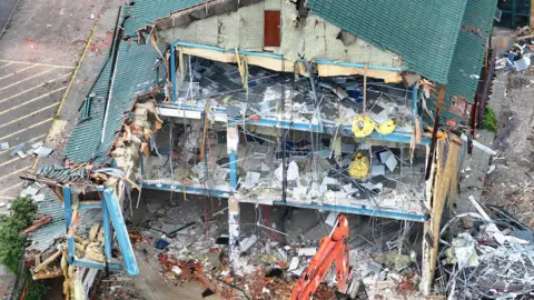Eddie Mitchell Aerial photograph of former Body Shop headquarters in Littlehampton being demolished by machinery. The green roof and one of the walls of the three-storey building have been torn down, exposing the insides. Construction rubble and metals scatter the floor and edges of the building, with an orange excavator at work at the bottom of the image.