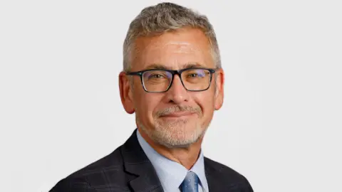 Prof Shane O'Neill is smiling in a university website picture. He has grey hair and beard and is wearing dark glasses, a dark jacket and grey shirt and tie