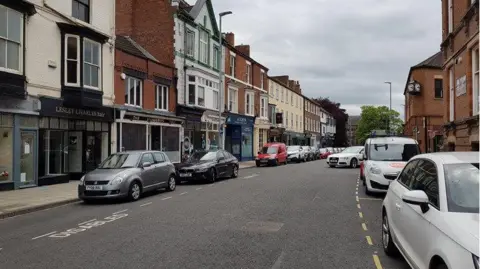 Road with cars parked on either side. There are shops on the road. 