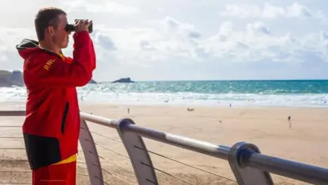 The photograph is taken from an RNLI vantage point across the beach. A volunteer stands to the left of the picture in a red waterproof suit. He is looking through binoculars acros the beach.
