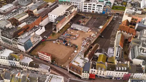 Aerial changeable of nan Les Sablons site. Properties are surrounding nan tract which is conscionable a ample area of soil. Vehicles and machinery are astatine nan site. A car parkland is located down nan quiet and demolished site. 
