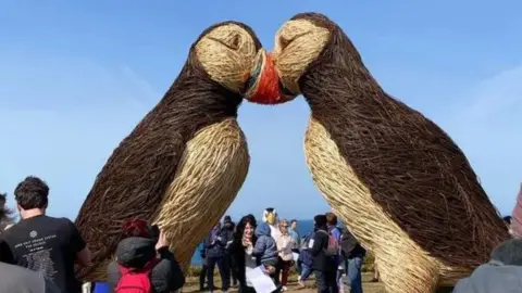 A wide, side shot of the two giant puffin sculptures with their heads together and crowds all around. The willow is not frayed as this was taken before the storm. A clear blue sky is visible in the background.